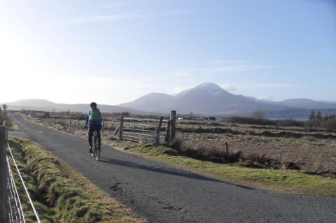 Photo showing person cycling on single track road with snow-capped mountain in the background